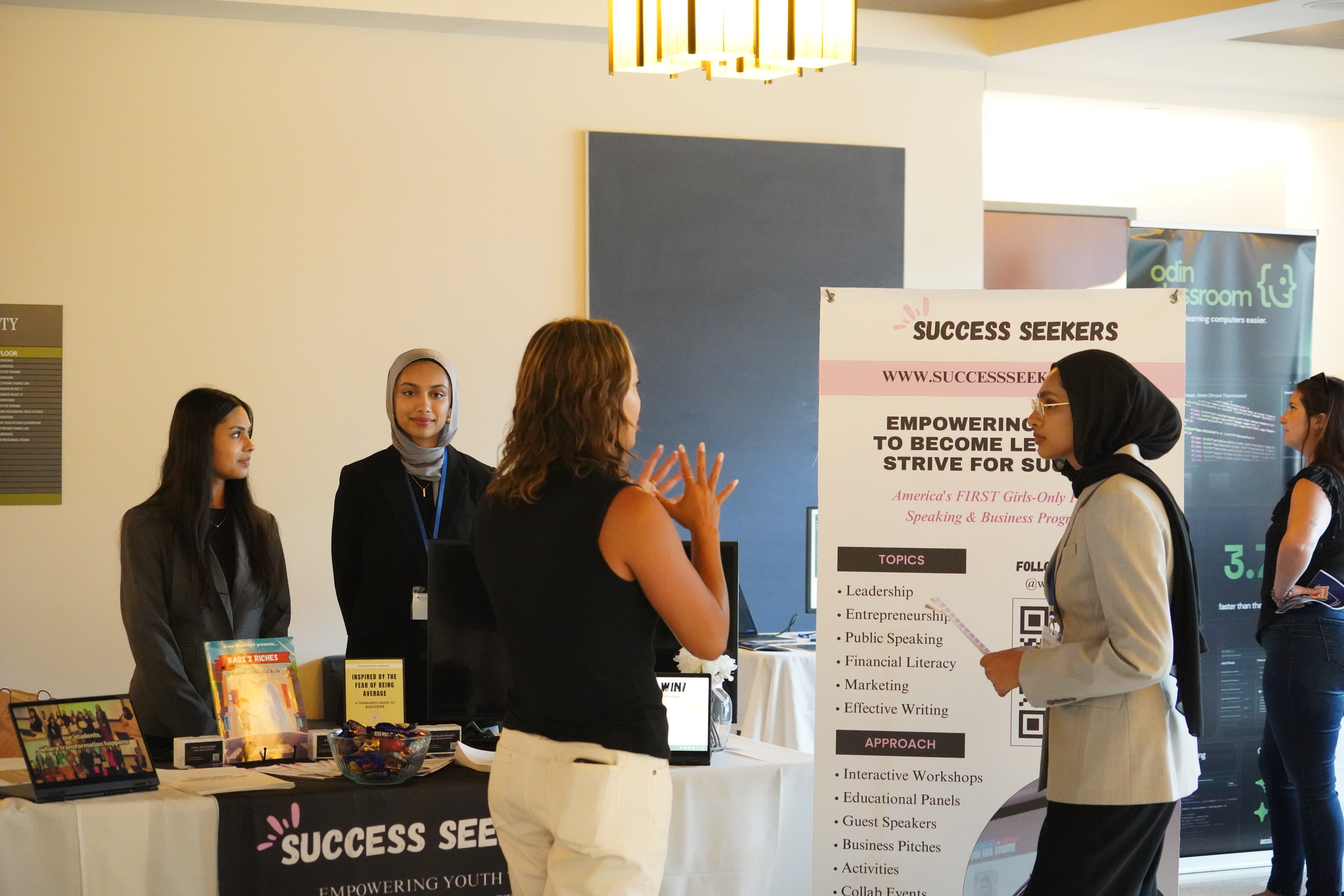 Two young women staff a table with a Success Seekers banner. They are mid-conversation with a table visitor